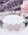 A Rose Quartz Gemstone stretch bracelet made with 2 pink rose quartz gemstones, translucent aura beads, and pink jasper gemstones sit on a white table. 