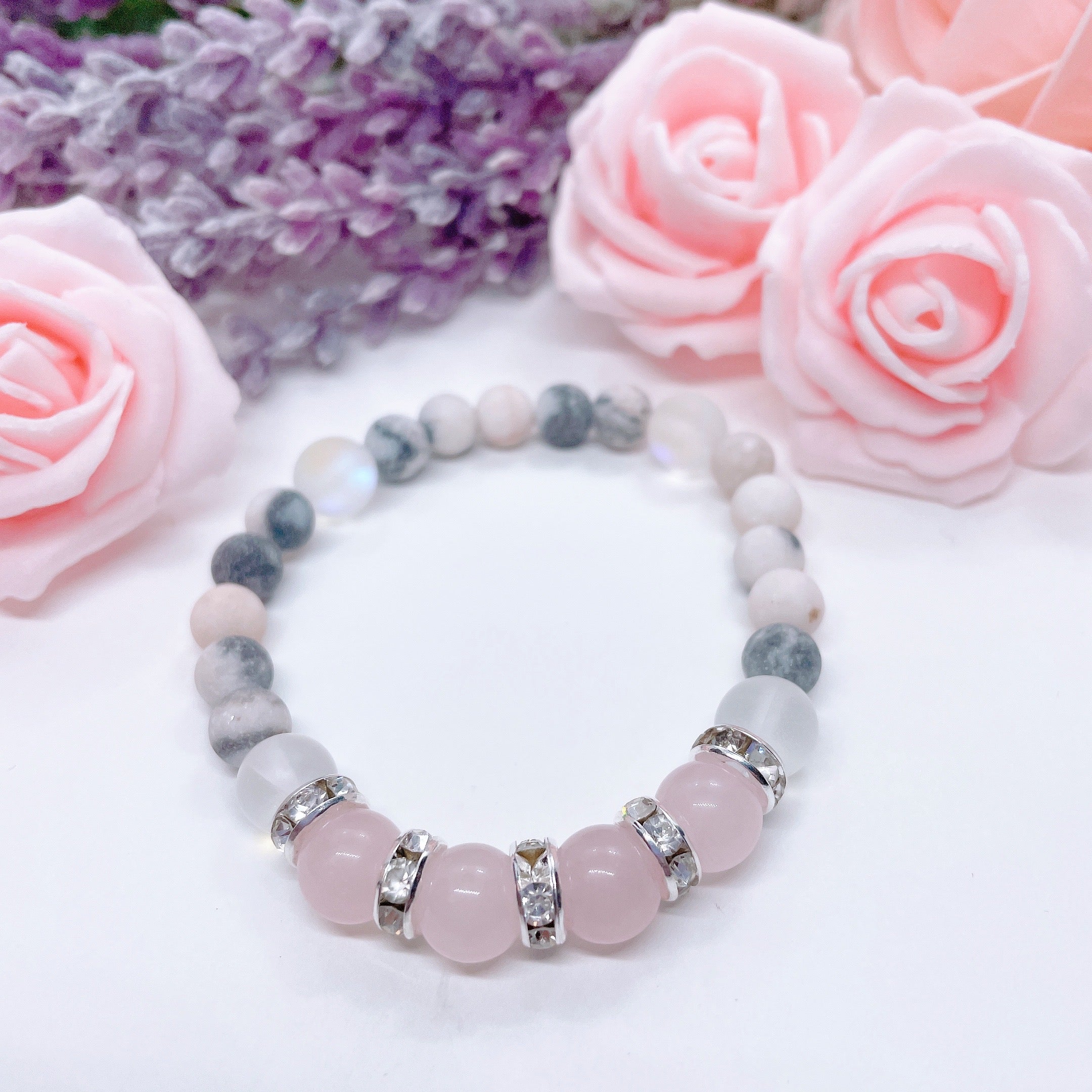 A Rose Quartz Rhinestone Companion Gemstone stretch bracelet made with 4 pink rose quartz gemstones, translucent aura beads, pink jasper gemstones, and rhinestone accents for added sparkle sits on a white table. 
